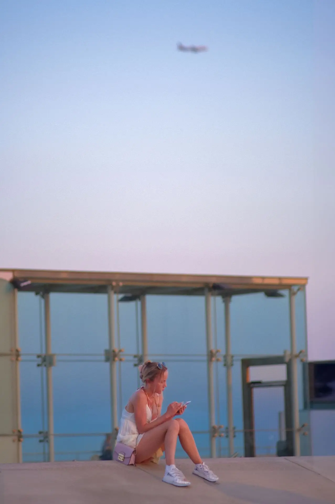 Sentada ante una vista infinita, una mujer parece absorta en su pantalla, mientras la arquitectura de vidrio refleja los suaves tonos del crepúsculo. Una escena que captura la soledad urbana y la belleza de los momentos efímeros en una ciudad donde cada detalle se convierte en una obra de arte.