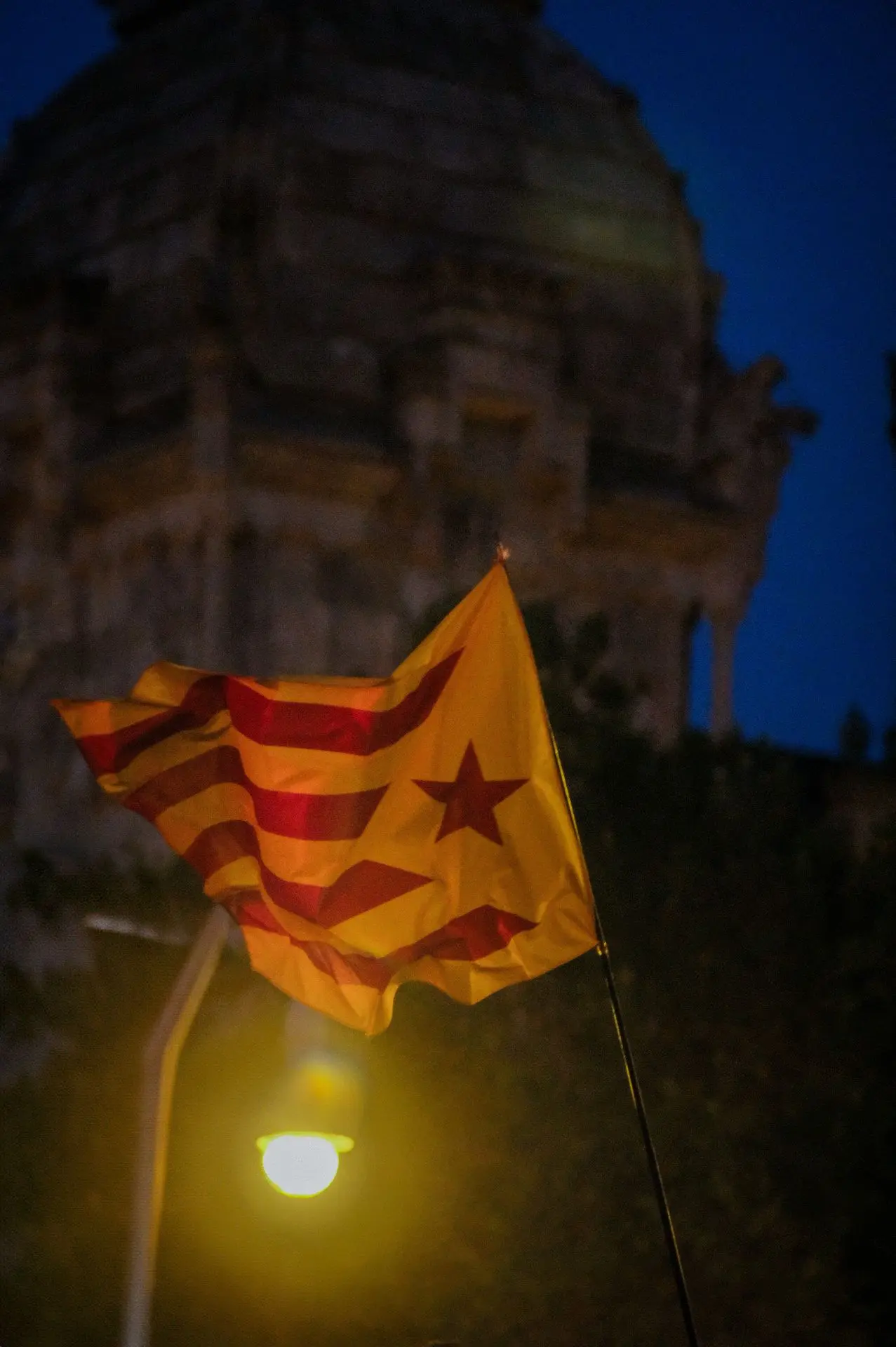 Esta imagen es una poderosa declaración visual de identidad y resistencia. La estelada, la bandera de la independencia catalana, ondea con dinamismo, simbolizando un movimiento profundamente arraigado en la historia y en constante evolución. La ausencia de la persona que la sostiene añade una capa de universalidad: esta bandera no representa solo a un individuo, sino a todo un colectivo, un sentimiento que trasciende un solo momento. La interacción entre la luz y el movimiento da a la tela una calidad escultórica, resaltando la fluidez de la expresión política y cultural en el cambiante paisaje de Barcelona.