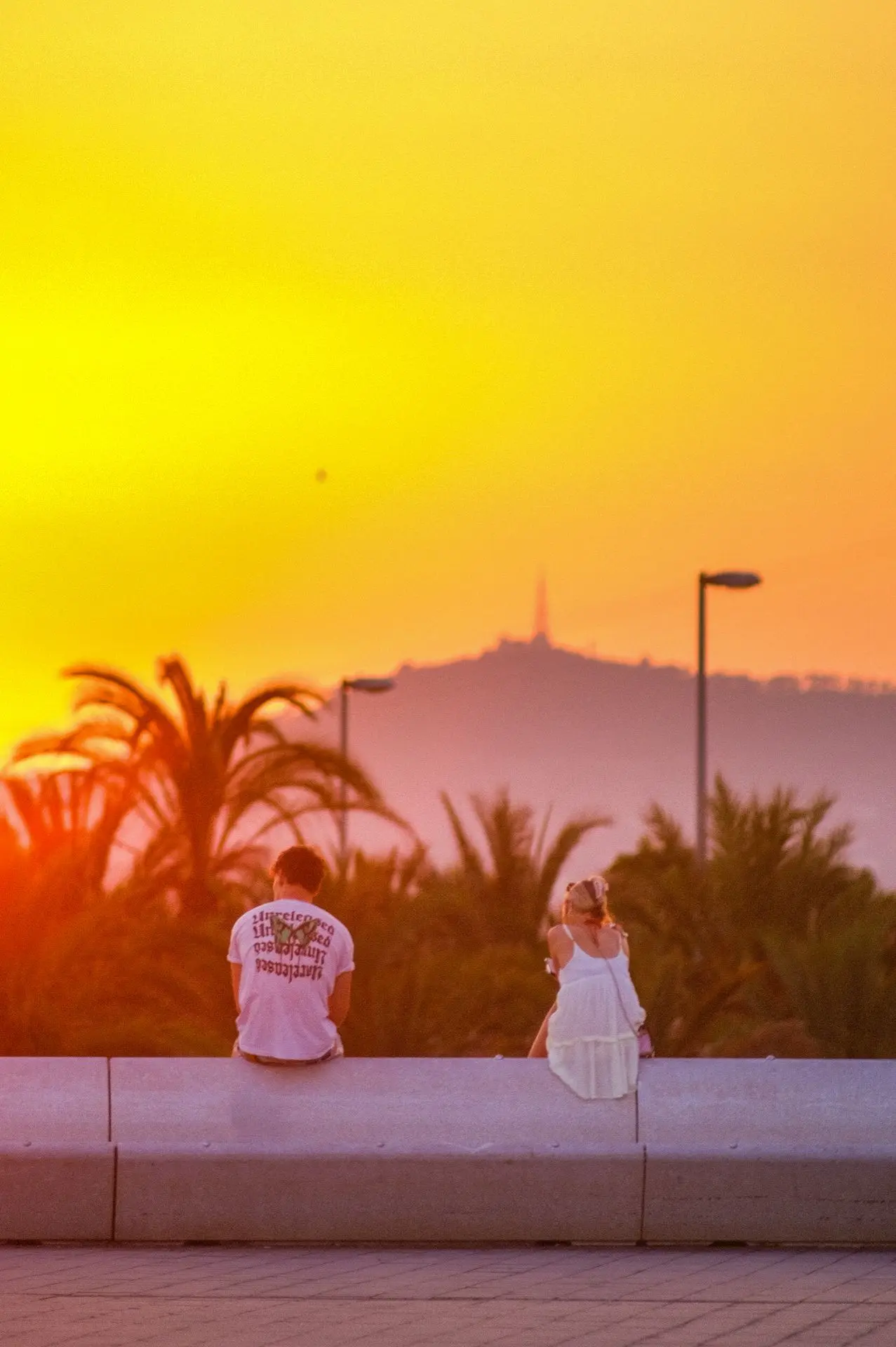 Una escena que captura la magia de un atardecer en Barcelona, donde dos siluetas contemplan el horizonte encendido. La atmósfera cálida y la profundidad de campo crean una composición visual poética, que encarna la esencia del viaje y la contemplación urbana.